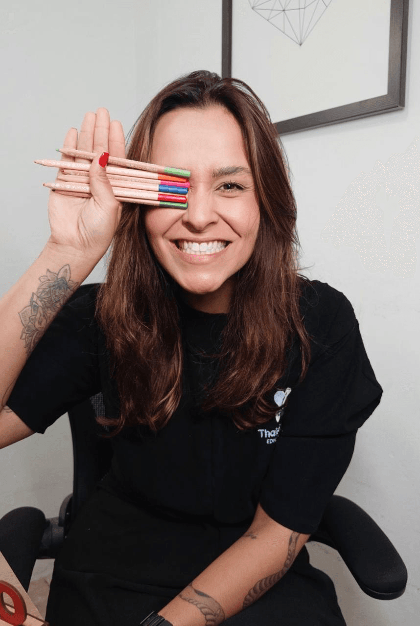 Thais Sampaio sorrindo, com lápis coloridos junto ao rosto